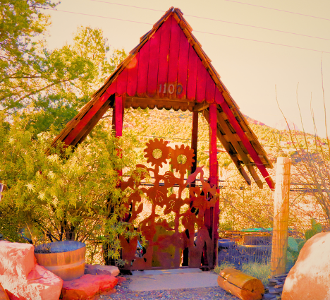 •Rustic red wooden entry gate at Apple Bob’s Cider Shack in Sedona, decorated with metal sunflower artwork and surrounded by desert plants